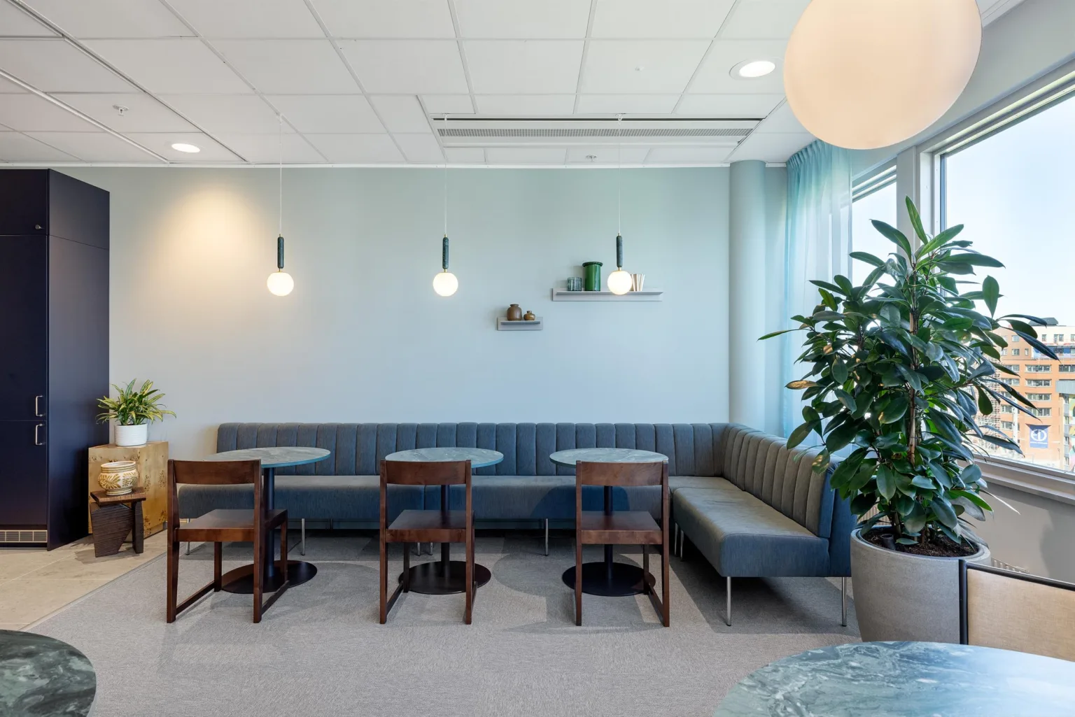 Ett modernt kaféområde med blå väggbänk, träbord och stolar, vägglampor samt växter på hyllor.