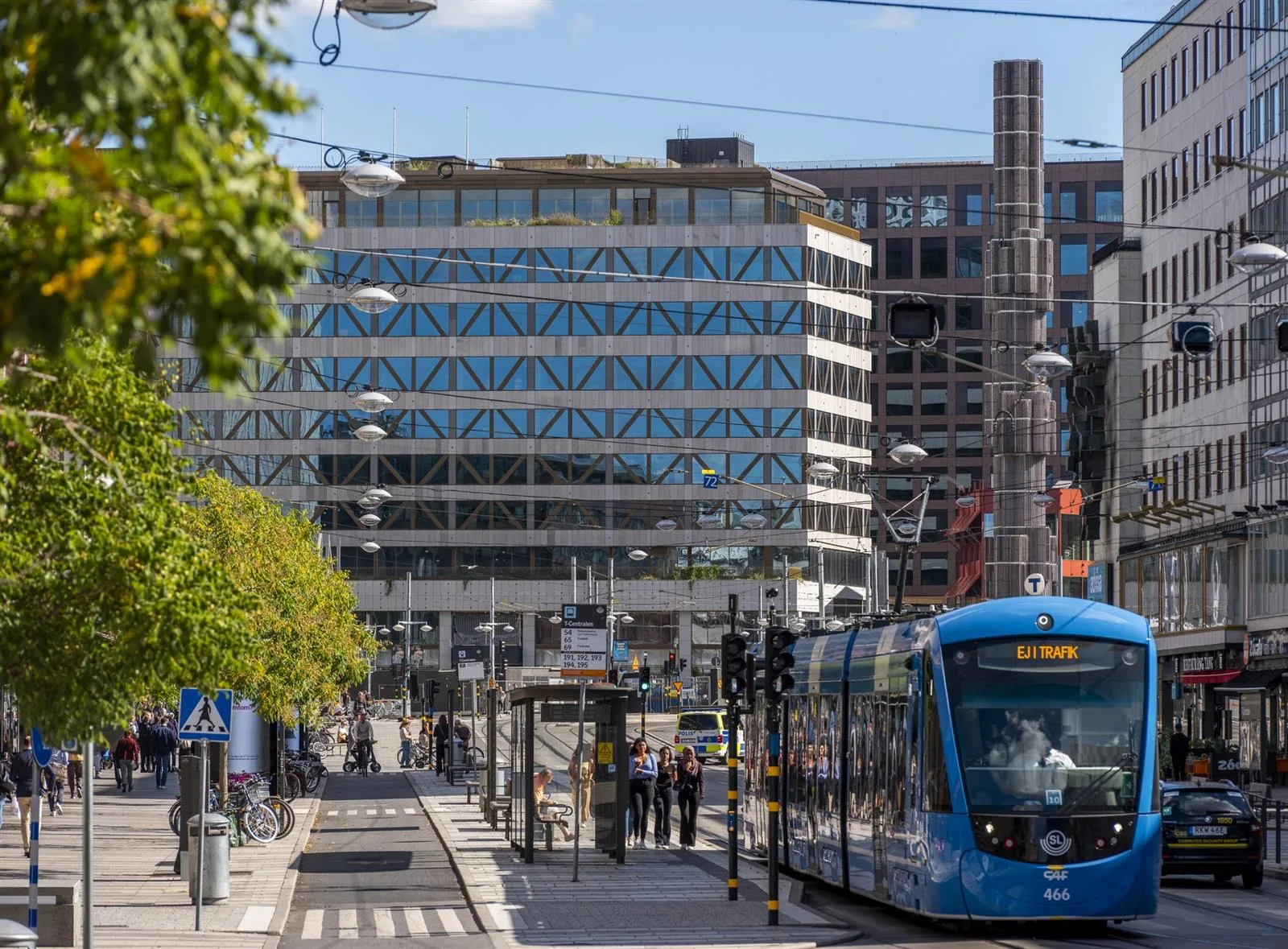 En blå spårvagn kör på en stadsgata framför en byggnad med geometriska fönster och människor vid en hållplats.