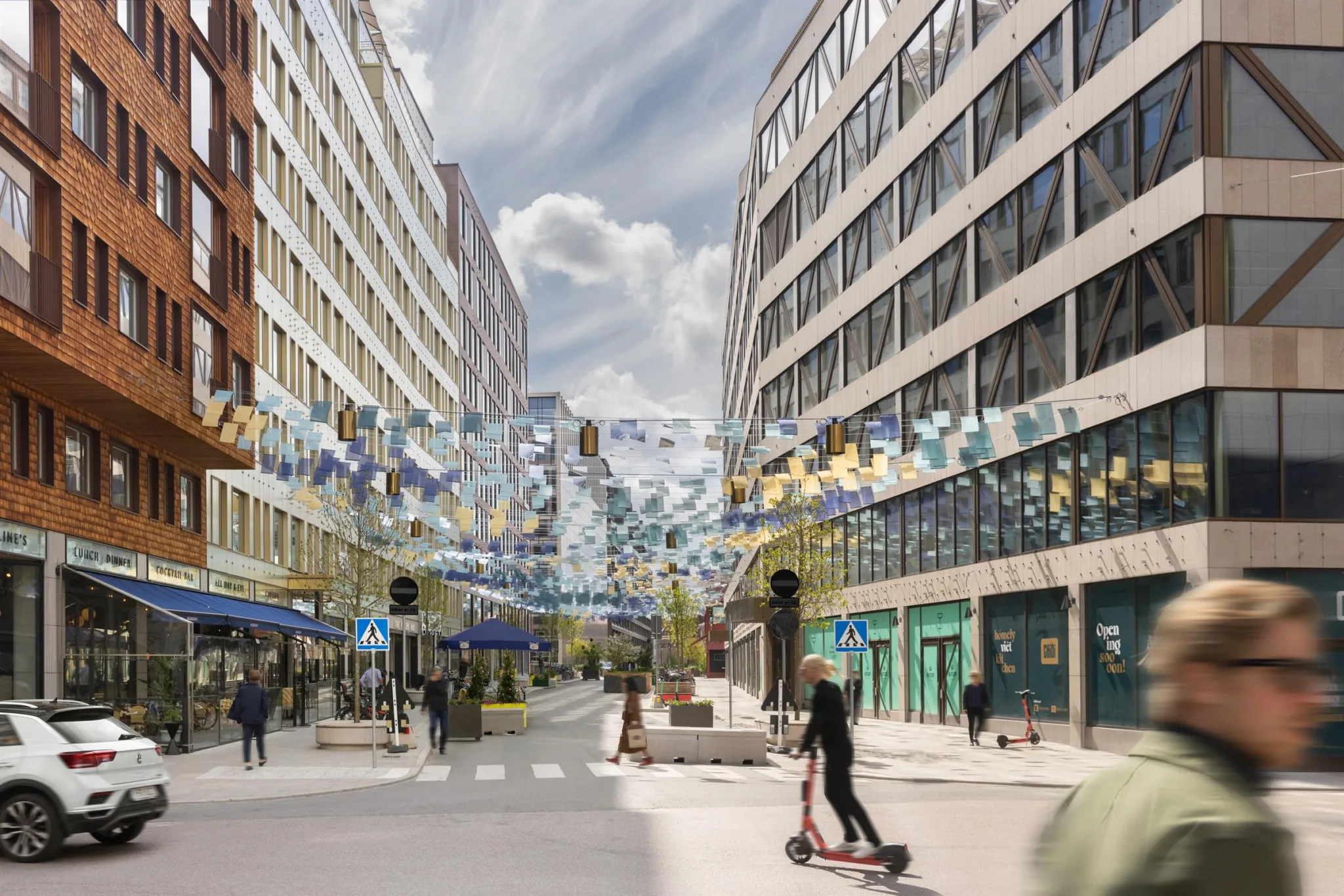 En stadsgata kantad av moderna byggnader med glasfasader, dekorerad med hängande färgglada band. En person åker elsparkcykel på den öppna trottoaren.