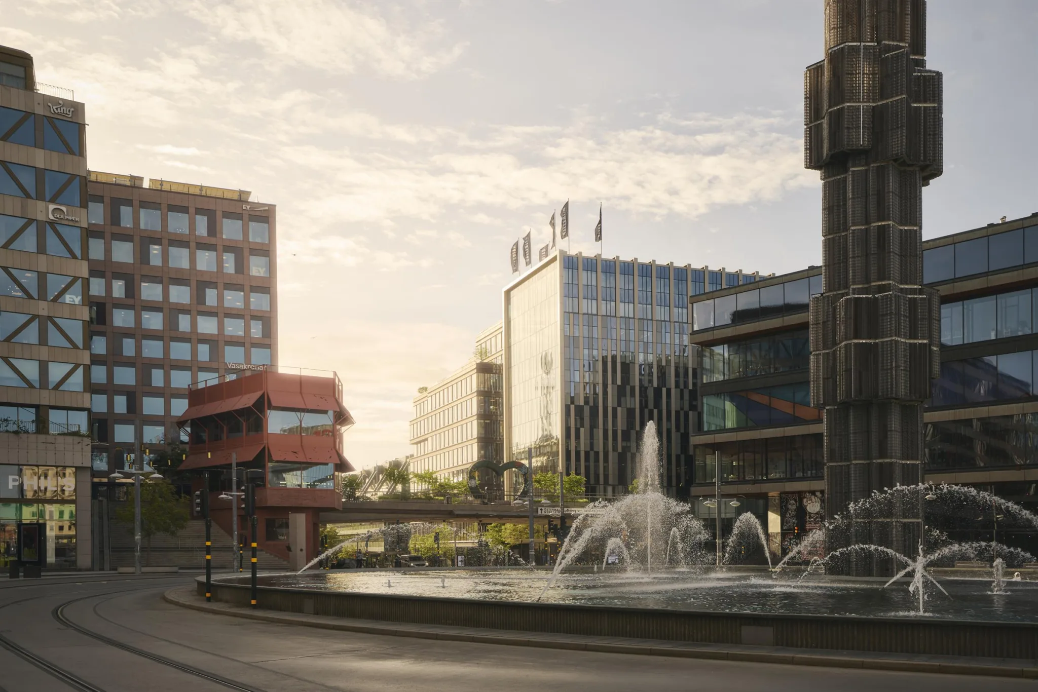En stadsmiljö med fontän och moderna byggnader, inklusive en röd takstruktur och hotellflaggor, under en ljus himmel.