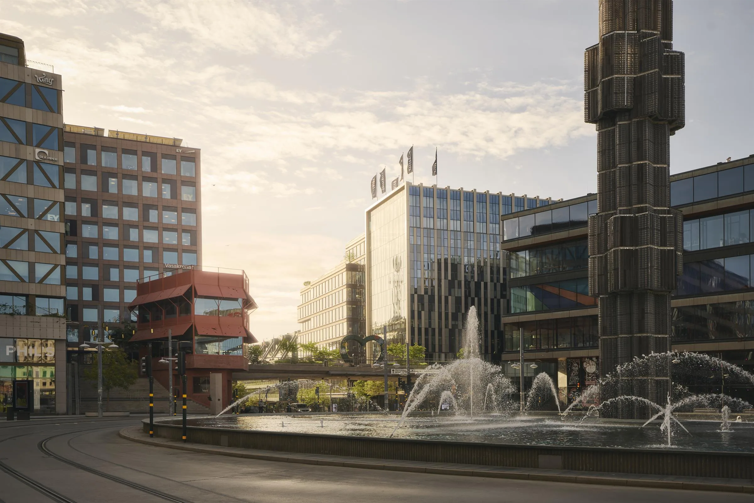 En stadsmiljö med fontän och moderna byggnader, inklusive en röd takstruktur och hotellflaggor, under en ljus himmel.
