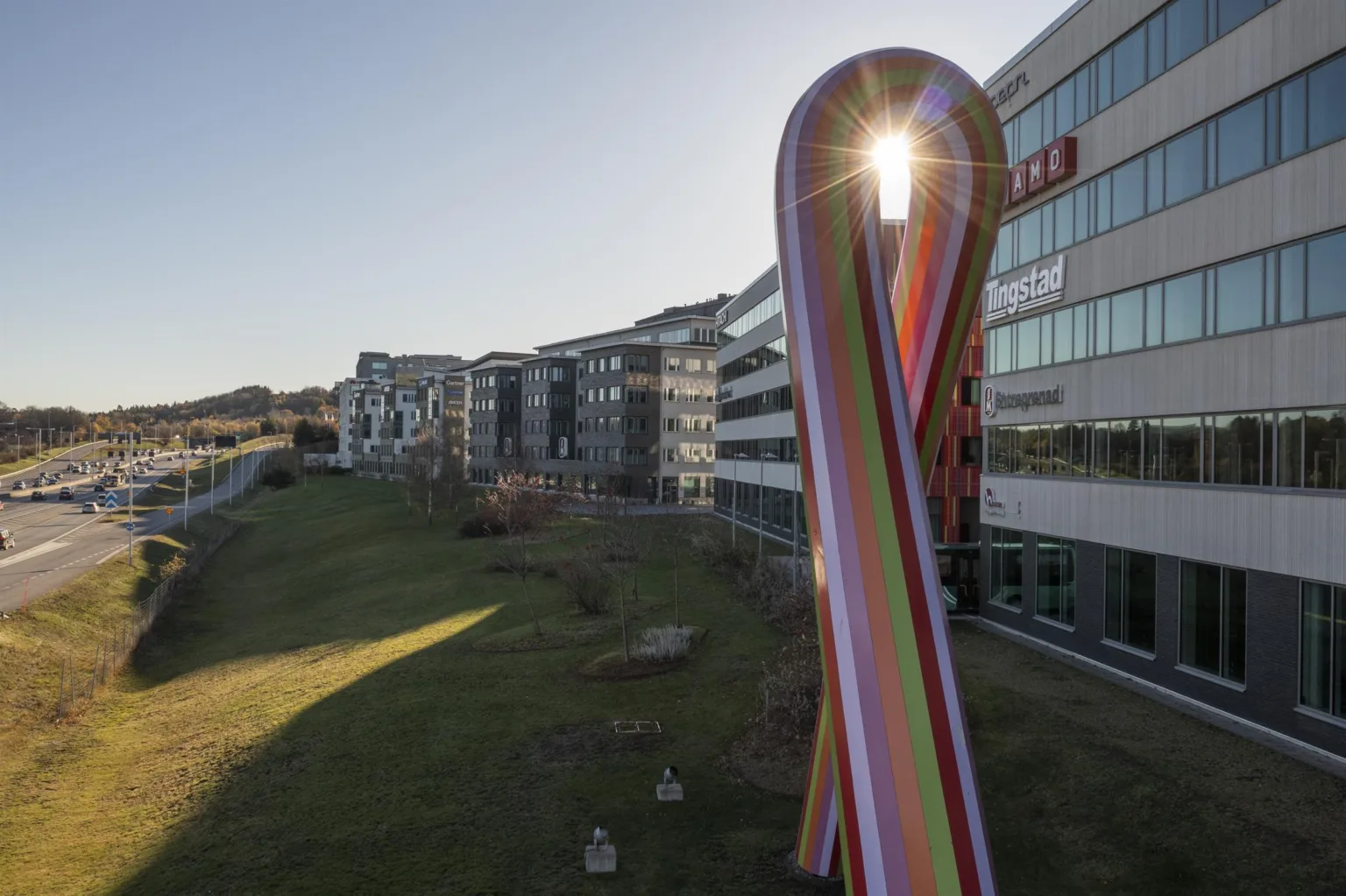 1. En stor färgglad skulptur i form av ett band står på en gräsmatta framför moderna kontorsbyggnader i dagsljus.