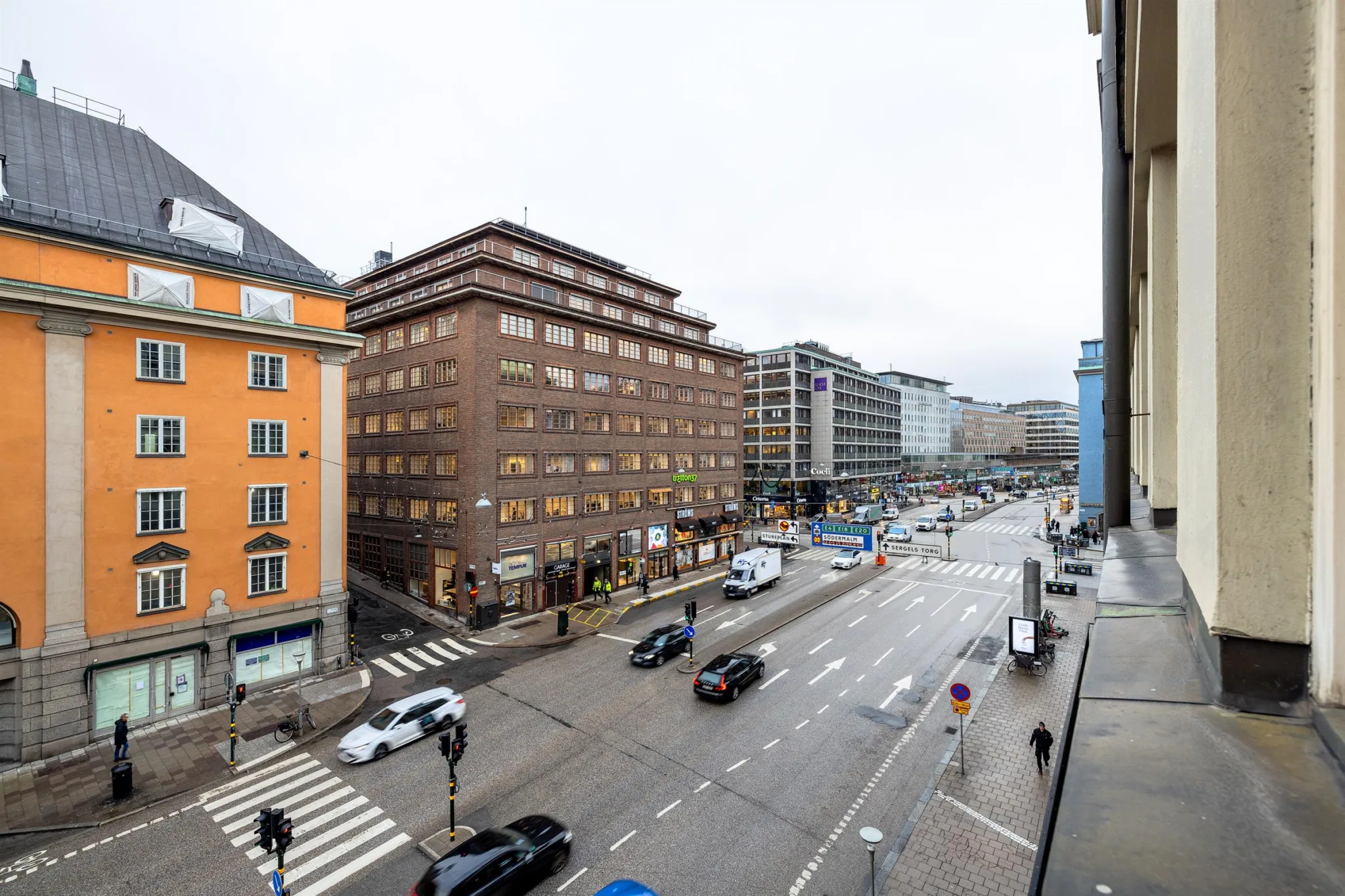 En stadsvy från ovan som visar en korsning med körande bilar, trottoarer och omgivande byggnader.
