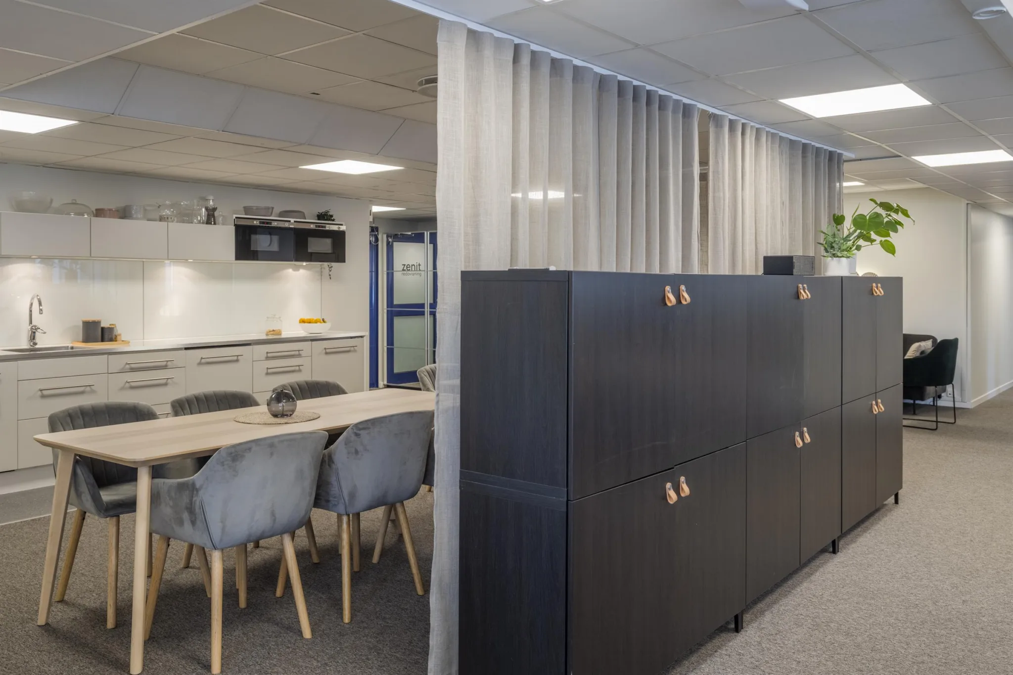 5. Kontorshörna med ett kök i bakgrunden, ett bord med grå stolar och en svart förvaringsmöbel med skjutdraperier bredvid.