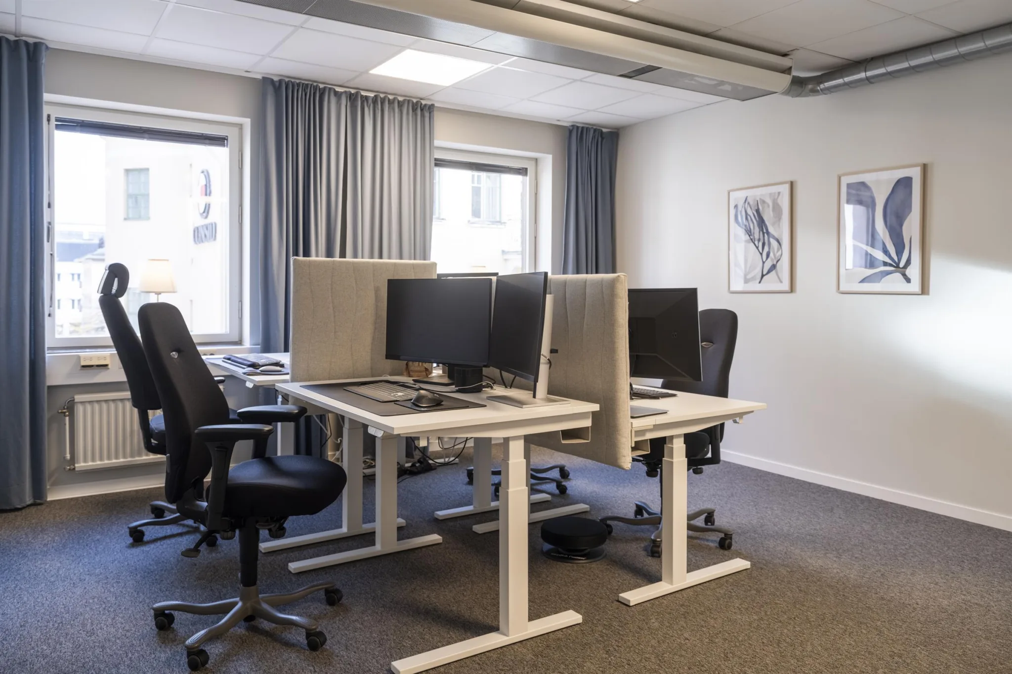 Ett kontorsutrymme med skrivbord, datorskärmar, kontorsstolar och fönster med gardiner.