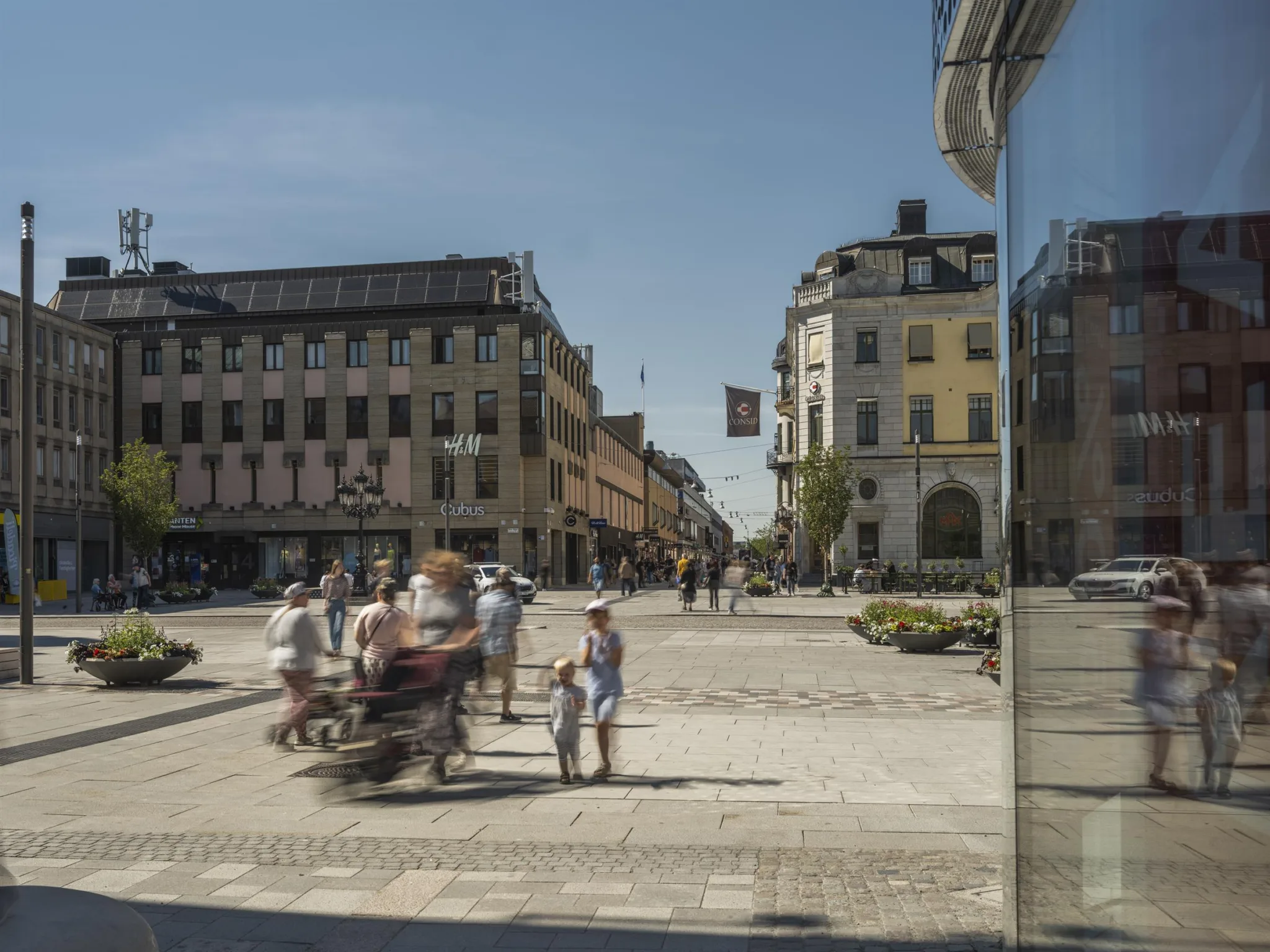 En stadsscen med fotgängare, cyklister och byggnader med klassisk arkitektur under en klarblå himmel.