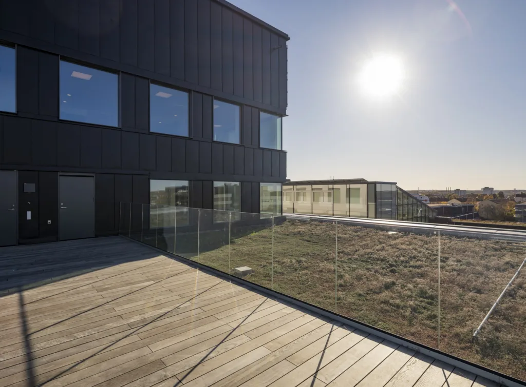 - En byggnad med mörk fasad och stora fönster, omgiven av en träterrass och glasräcke under en solig himmel.