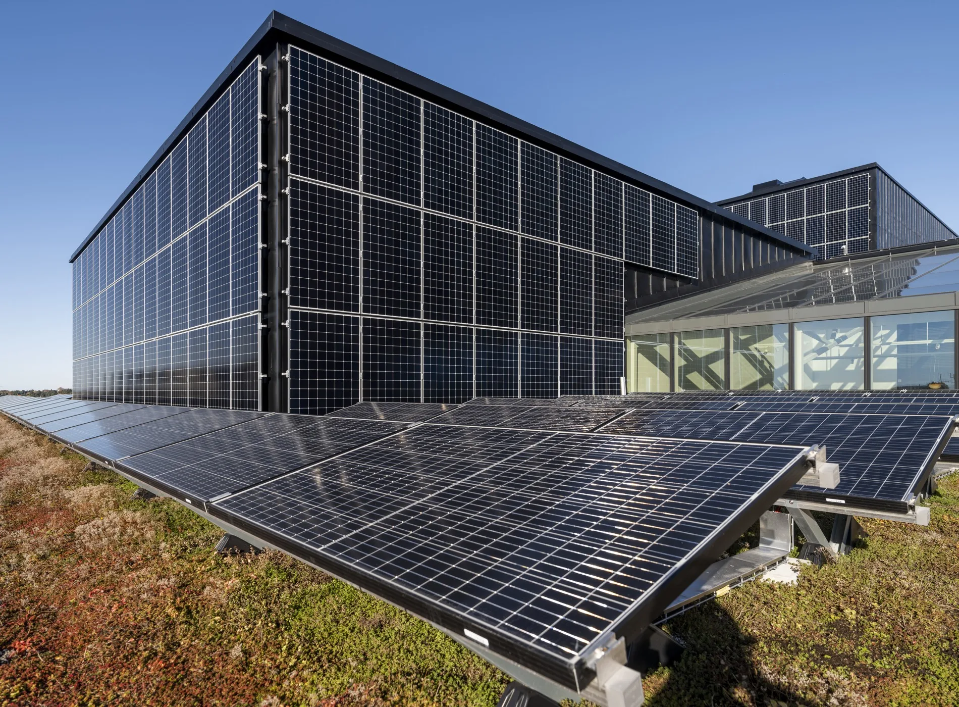 - Solpaneler monterade på tak och fasad av en modern byggnad, med klarblå himmel i bakgrunden.