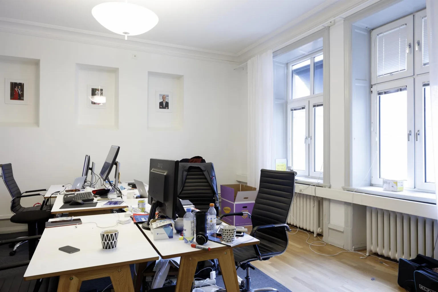 3. Ett kontor med skrivbord, datorskärmar och en ljus vägg med två fönster.