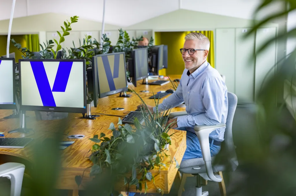 En person som sitter vid ett skrivbord med flera datorskärmar, omgiven av gröna växter, och ler mot kameran.