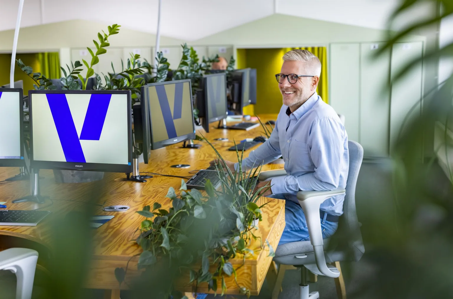 En person som sitter vid ett skrivbord med flera datorskärmar, omgiven av gröna växter, och ler mot kameran.