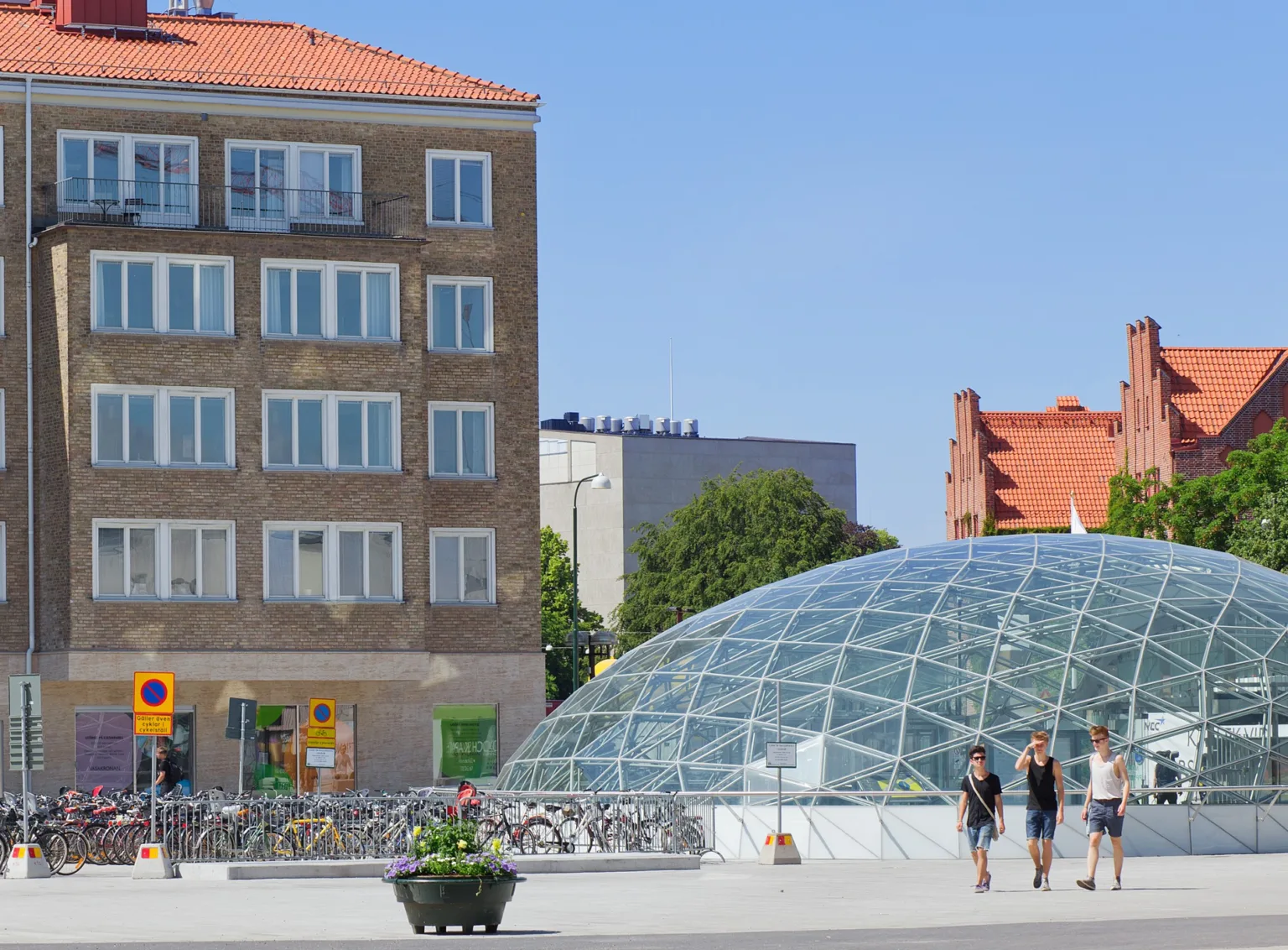 1. Ett glashus i modern design bredvid ett klassiskt flerfamiljshus, med människor som promenerar i förgrunden under en klarblå himmel.