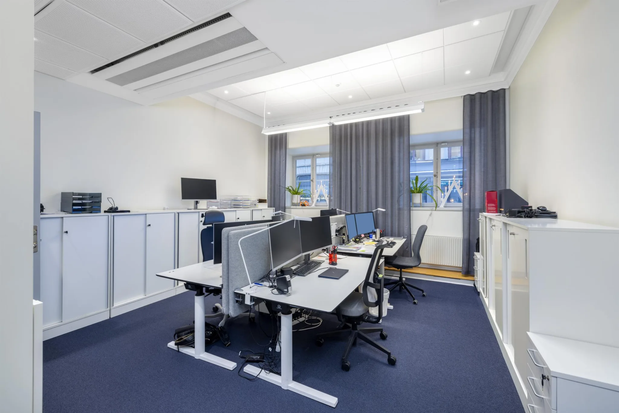Ett öppet kontorslandskap med flera arbetsplatser, skrivbord, datorer och stora fönster med gardiner.