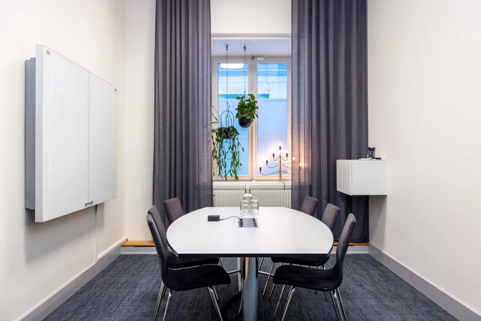 Ett mötesrum med ett vitt bord, svarta stolar, ett fönster med grå gardiner och växter på fönsterbrädan.