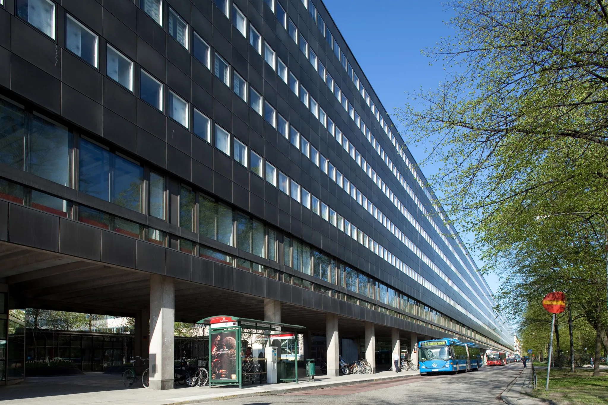En lång byggnad med svart fasad och stora fönster, en buss och telefonkiosk synliga vid trottoaren under en klar blå himmel.
