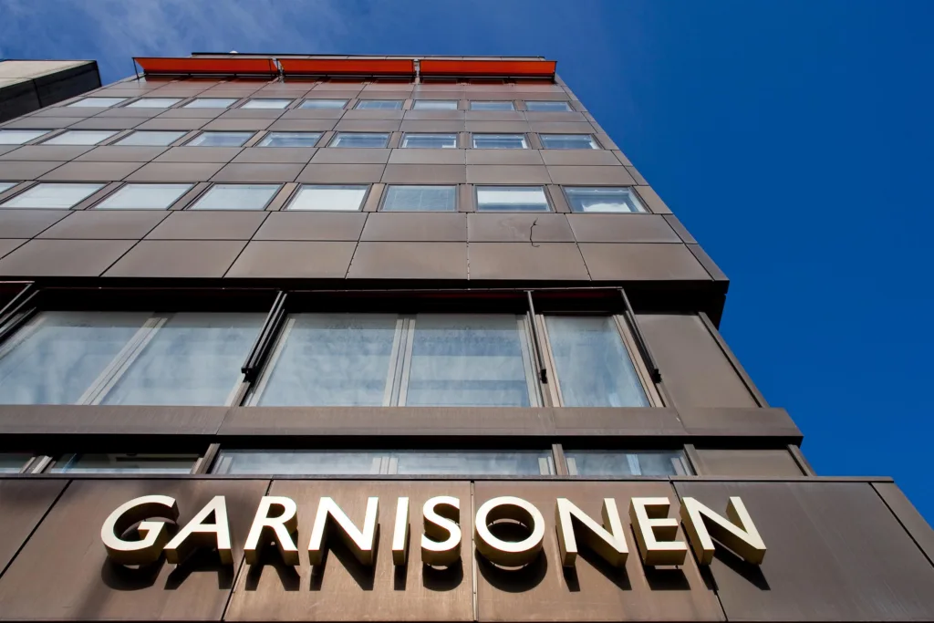 Byggnad med texten "Garnisonen" på fasaden, fotograferad från marknivå mot en blå himmel.