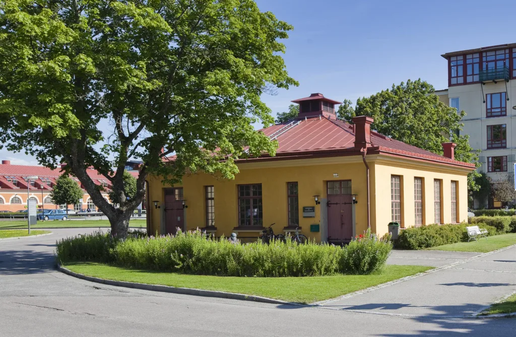En gul byggnad med röda tak, stora fönster och grönska framför, omgiven av träd och blå himmel.