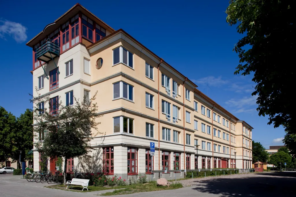 En beige flerfamiljsbyggnad med röda dörrar och fönster, omgiven av träd och en blå himmel.