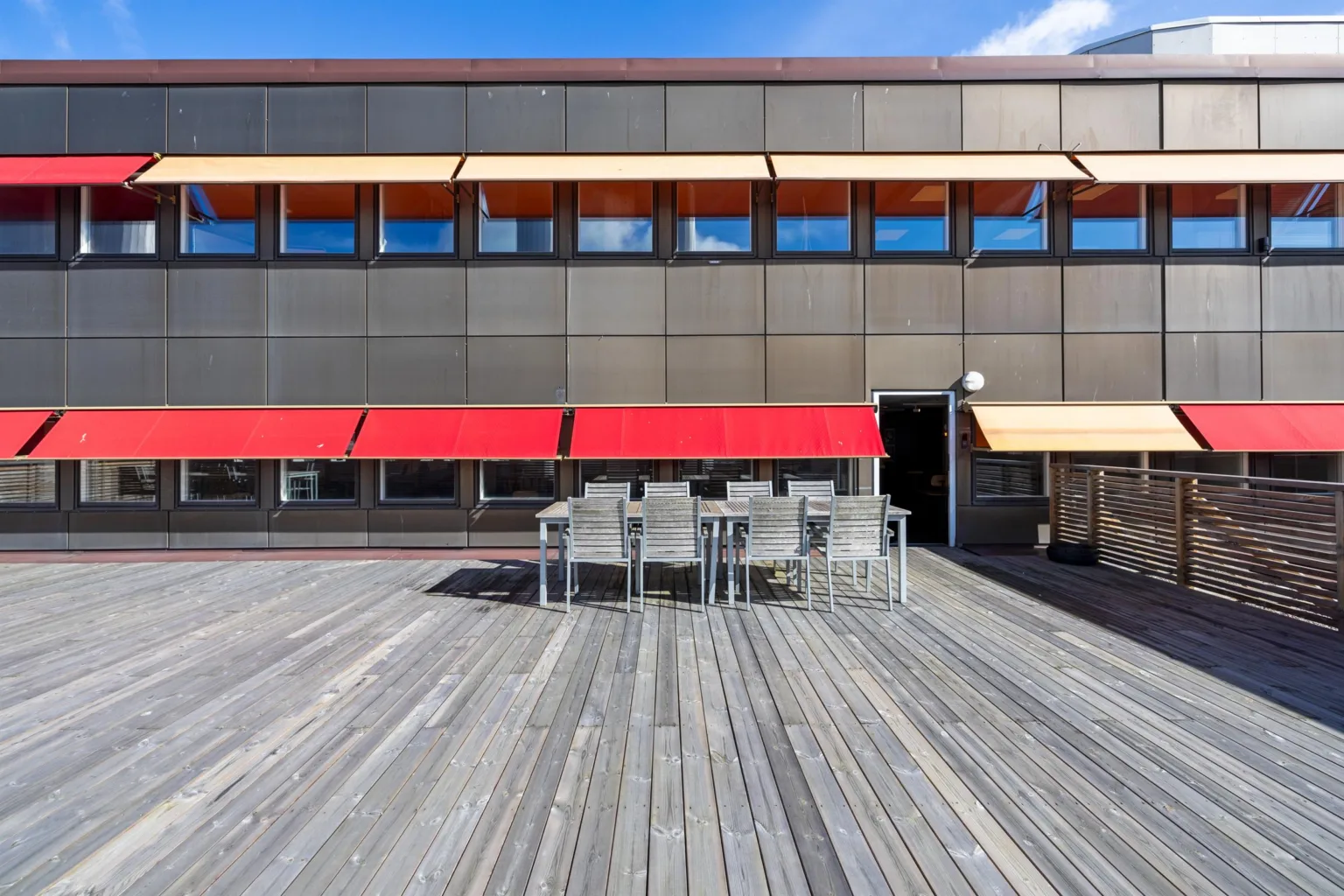En stor träbelagd terrass med röda markiser och utemöbler framför en modern byggnad.
