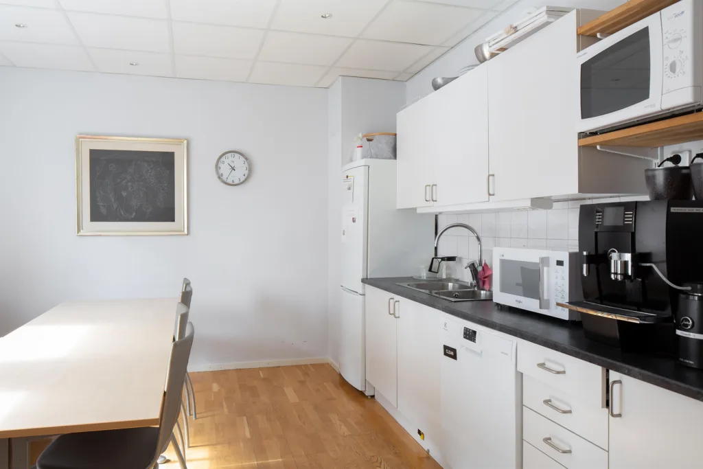 Ett ljust kök med vita skåp, en mikrovågsugn, en klocka på väggen och ett matbord.