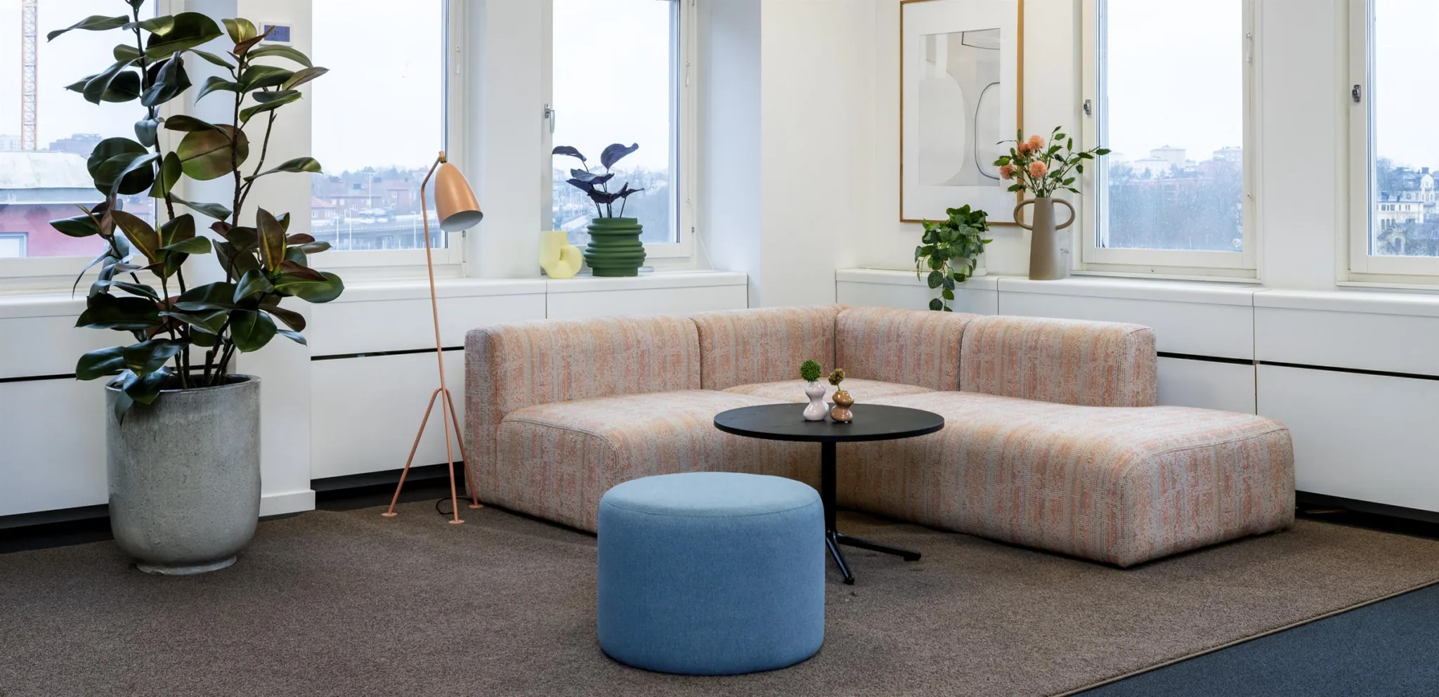 En mysig sittgrupp med en ljus hörnsoffa, ett runt soffbord, blå sittpuff och dekorativa växter.