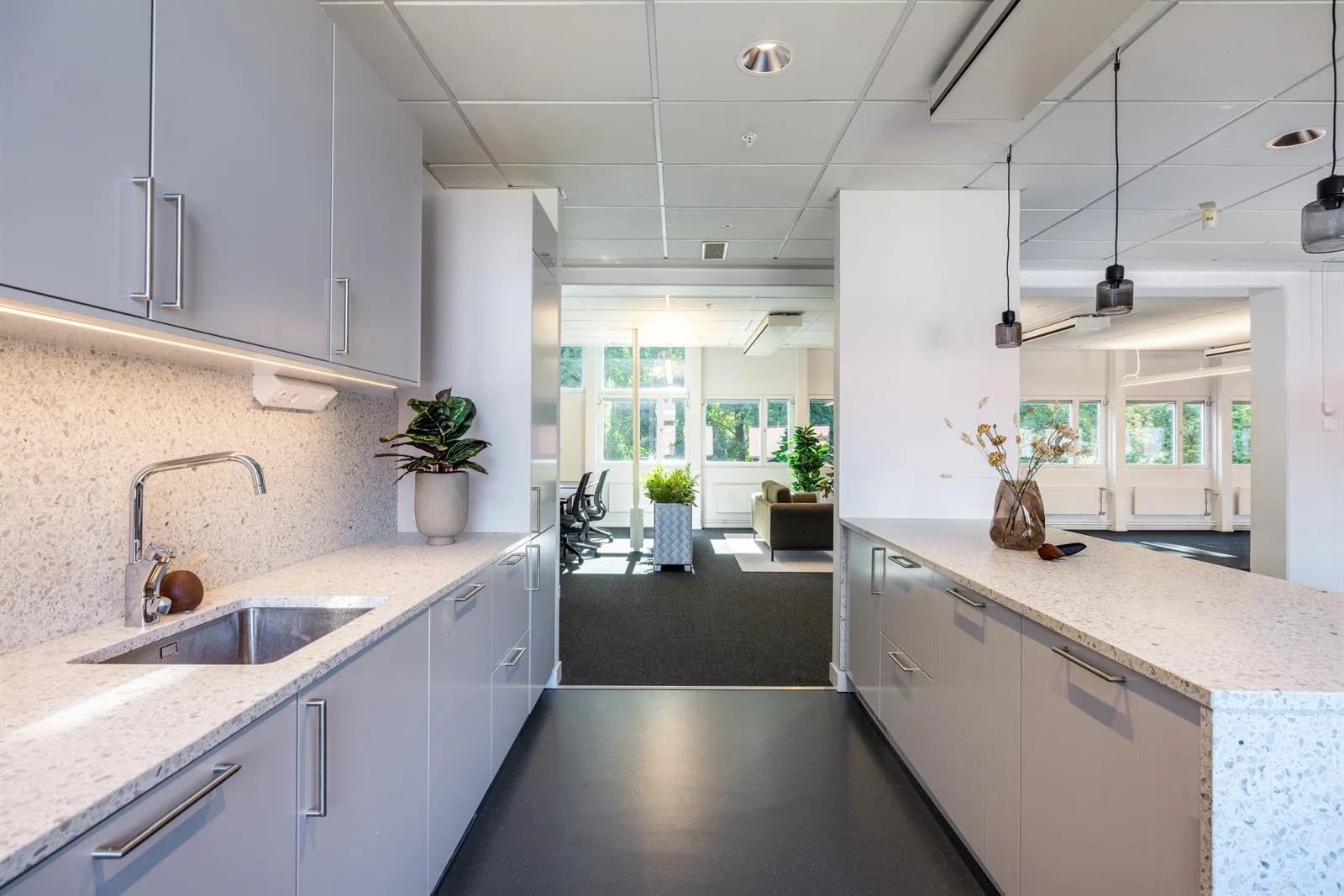 4. En ljus köksyta med vita skåp, diskbänk och växter, med utsikt mot ett kontorsutrymme i bakgrunden.