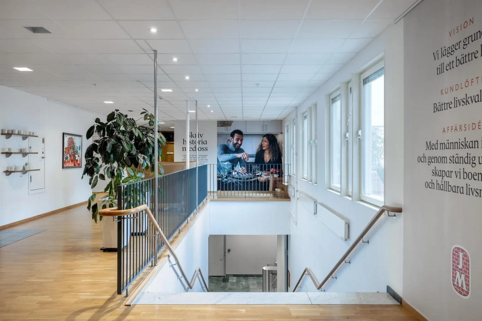5. En ljus trappa med glasräcke, stora fönster och en vägg med en tavla föreställande två personer.