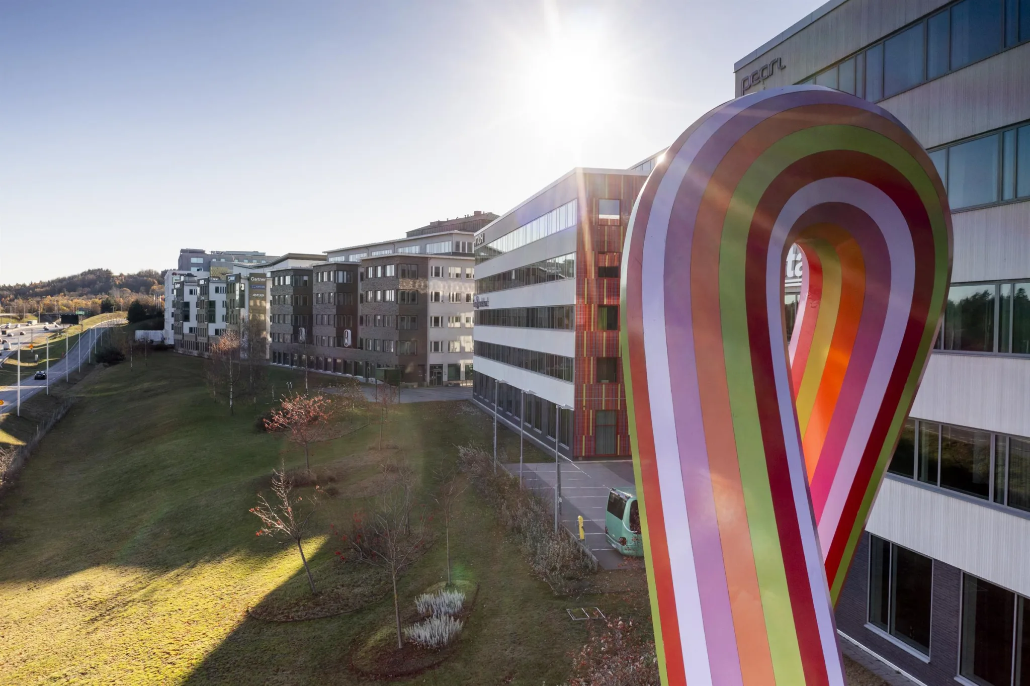Färgglad konstskulptur i förgrunden med moderna byggnader och en solig park i bakgrunden.