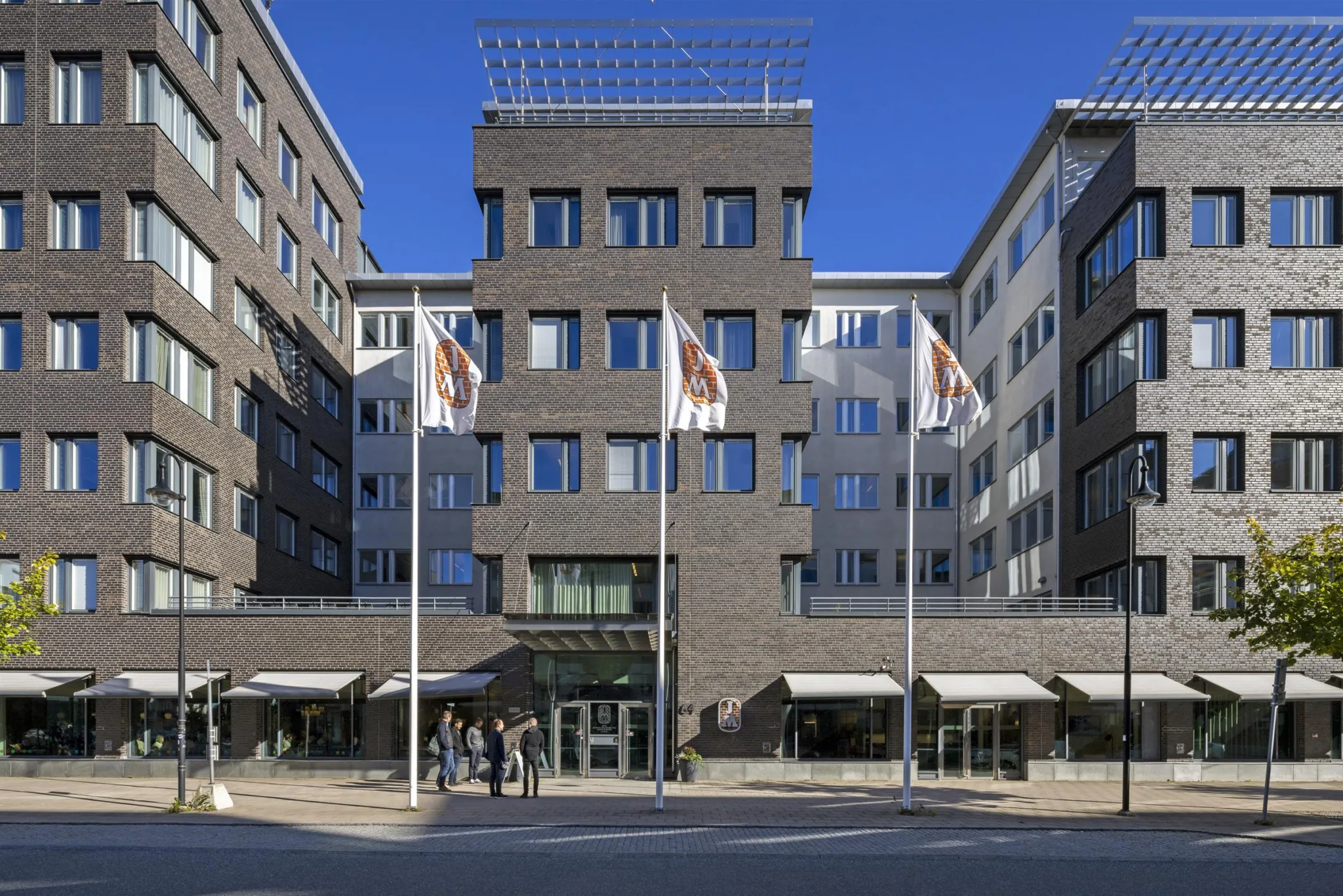En byggnad med mörka fasader, tre flaggor i förgrunden och ingång med glasdörrar.