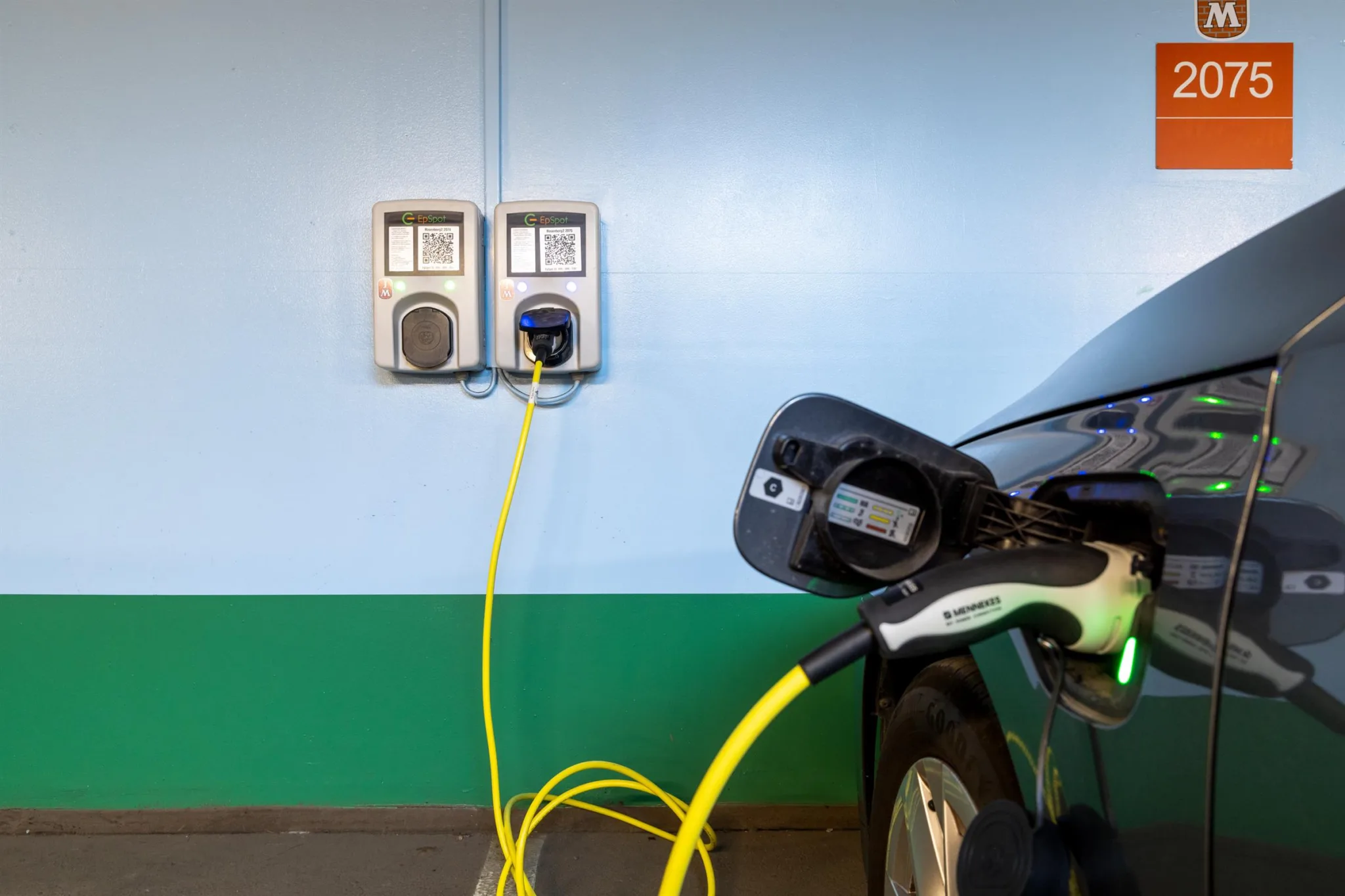 En elbil laddas med en gul kabel ansluten till en laddstation på en vägg med blå och grön färg.