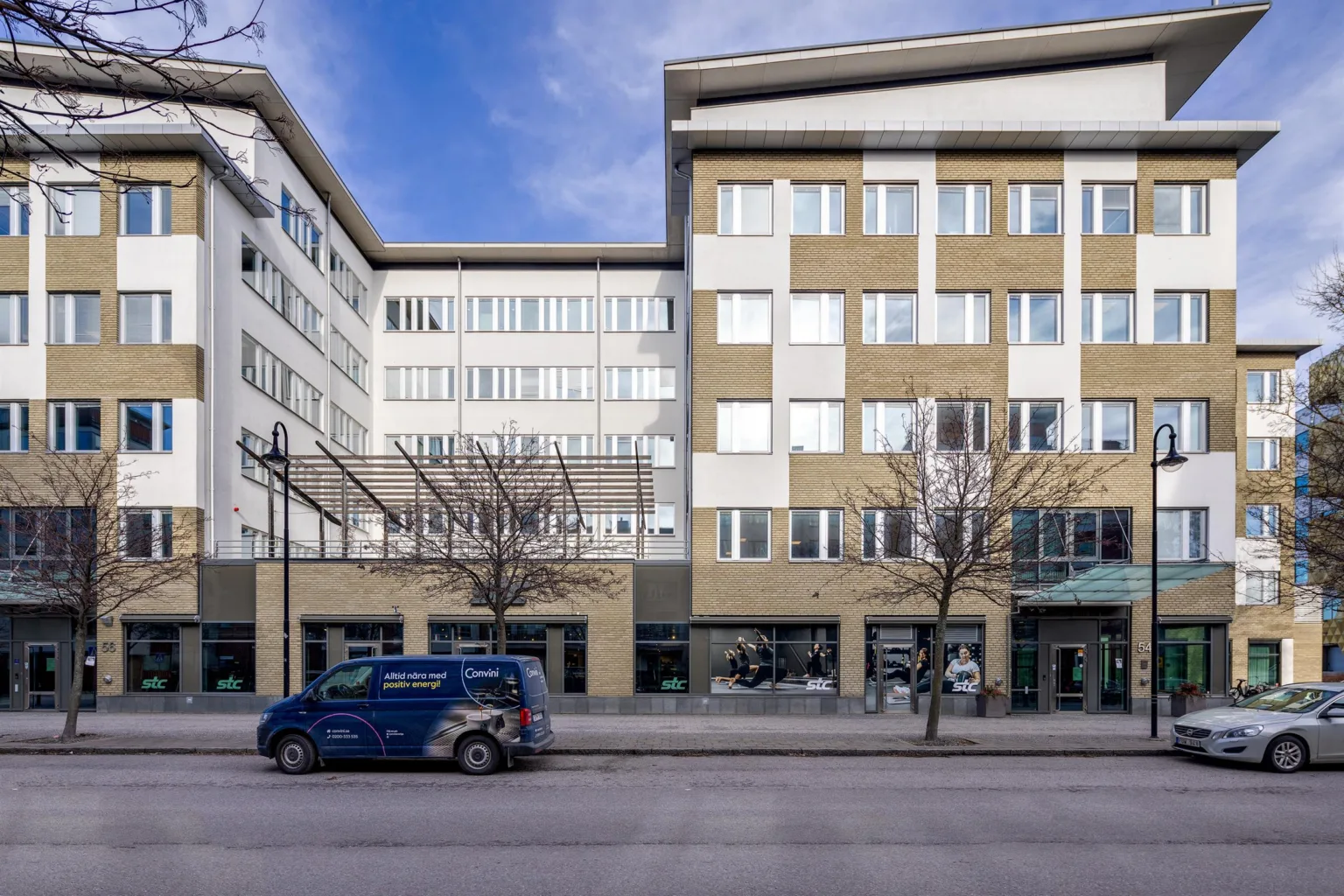 En byggnad med flera våningar, stora fönster och en blå bil parkerad på gatan framför.