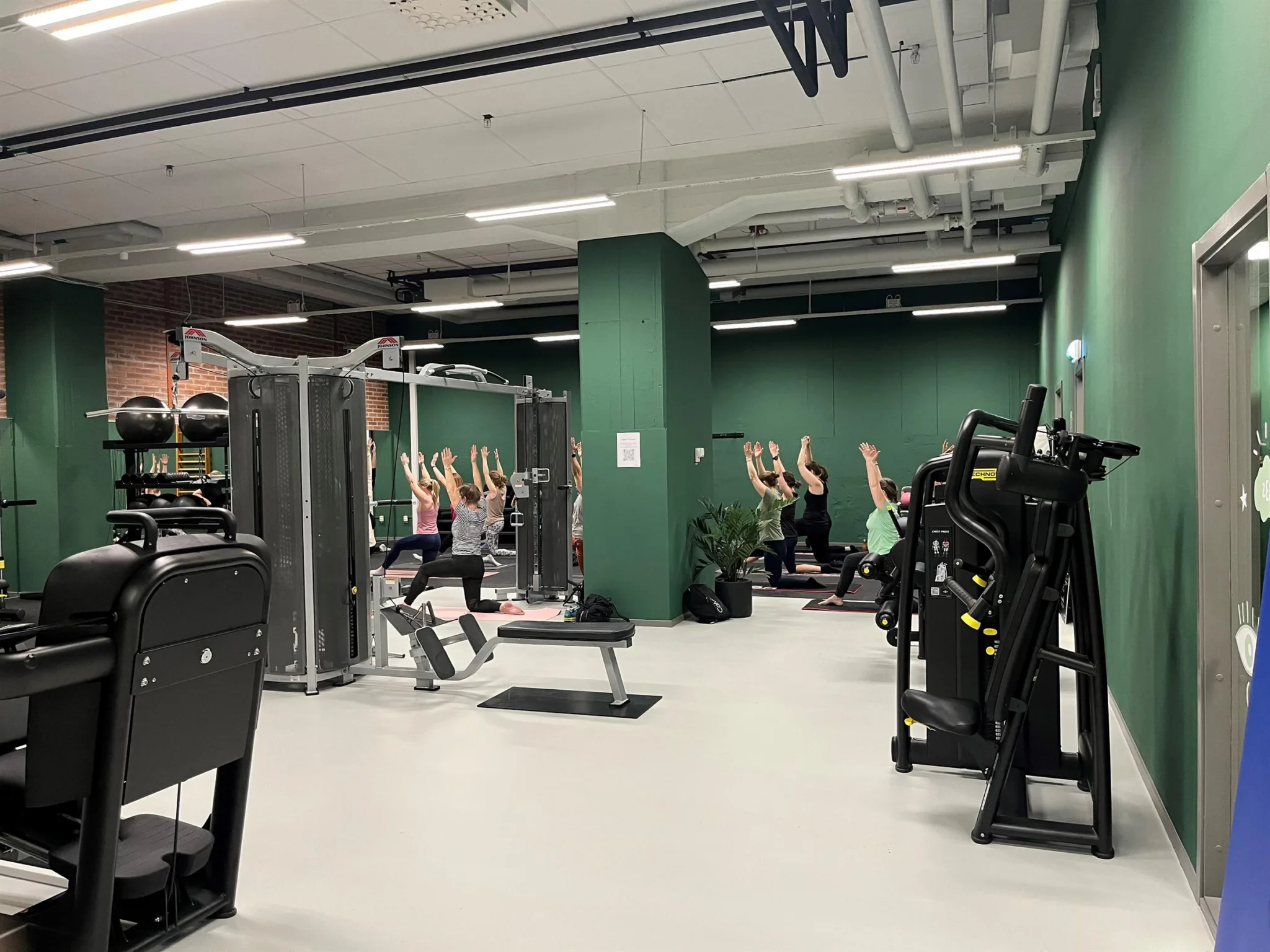Ett gym med träningsutrustning och människor som gör yoga i en sal med gröna väggar.