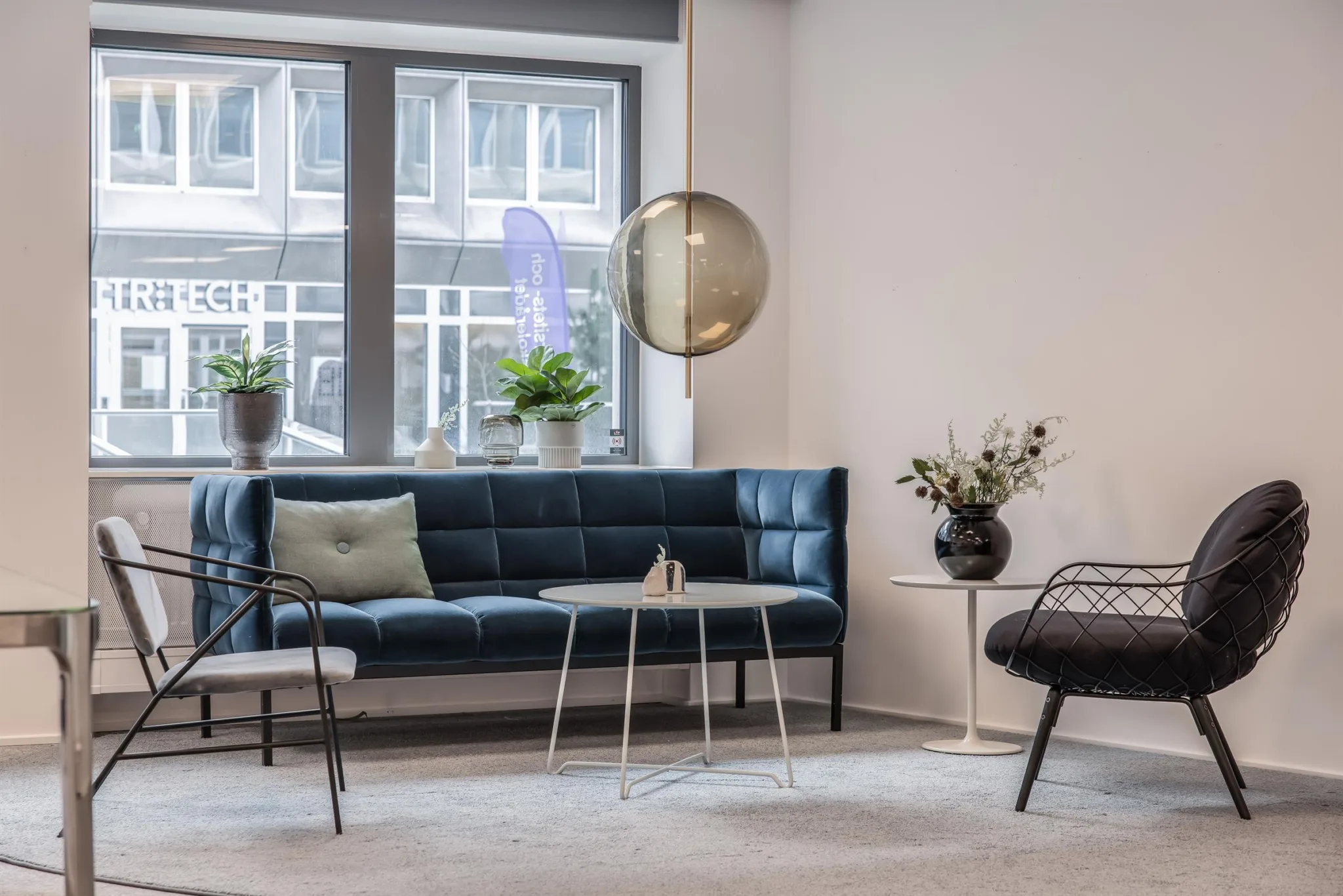 Ljus lounge med blå soffa, bord, stolar och dekorativa växter vid ett stort fönster.