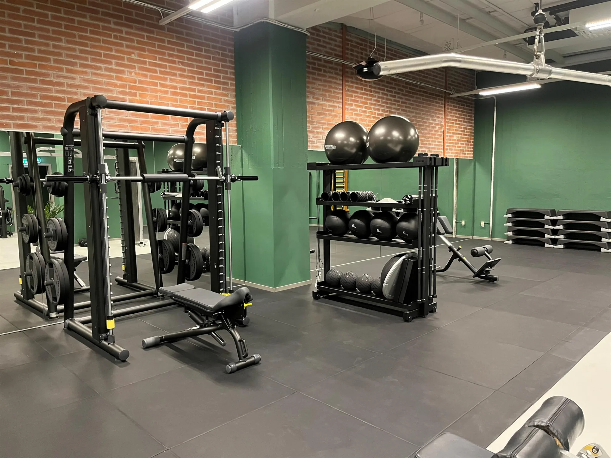 Ett gymrum med vikter, träningsmaskiner, stora bollar och gröna väggar med en tegelvägg i bakgrunden.