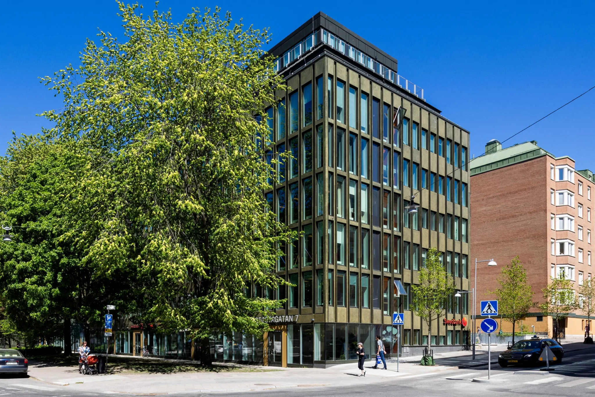Hörnvy av en modern byggnad med glasfönster, omgiven av träd och en blå himmel.