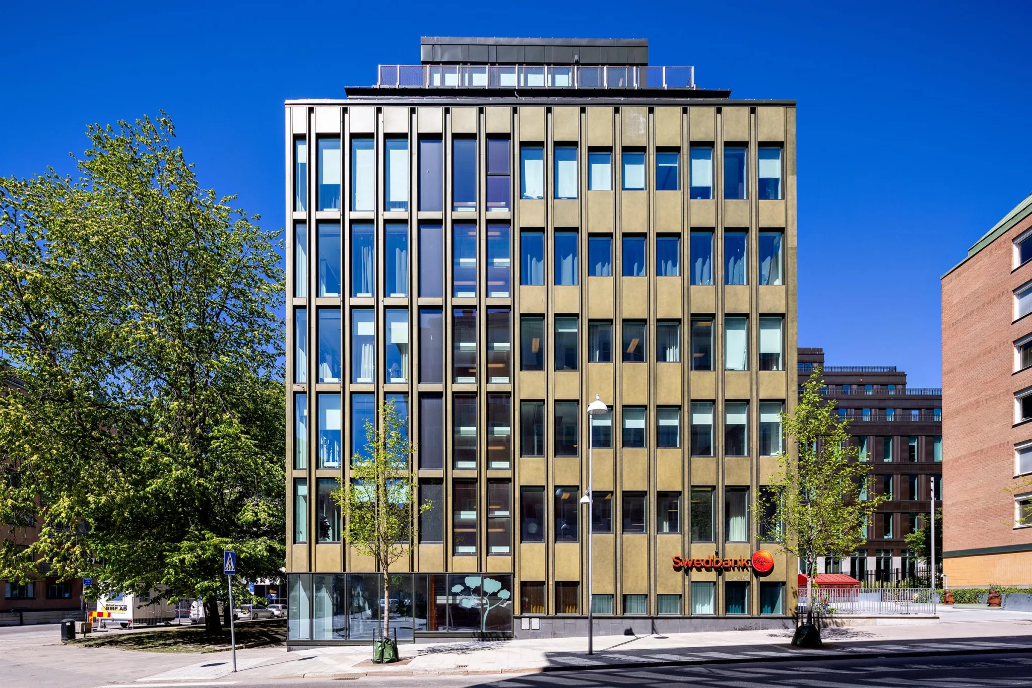 En modern byggnad med glasfasad och guldfärgade detaljer, omgiven av träd och en klar blå himmel.