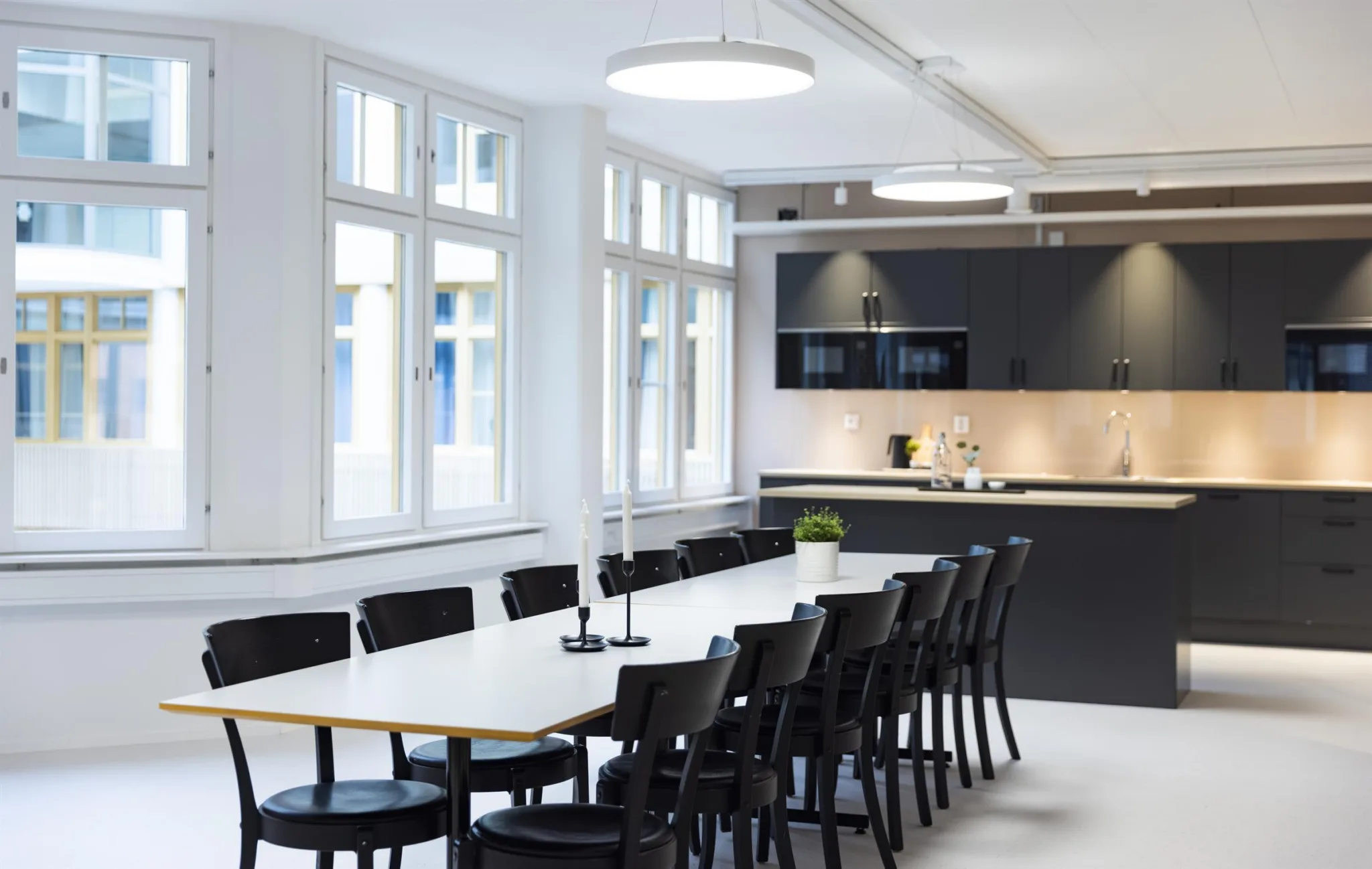 En matsal med ett långt vitt bord, svarta stolar och ett modernt kök i bakgrunden med stora fönster som släpper in ljus.