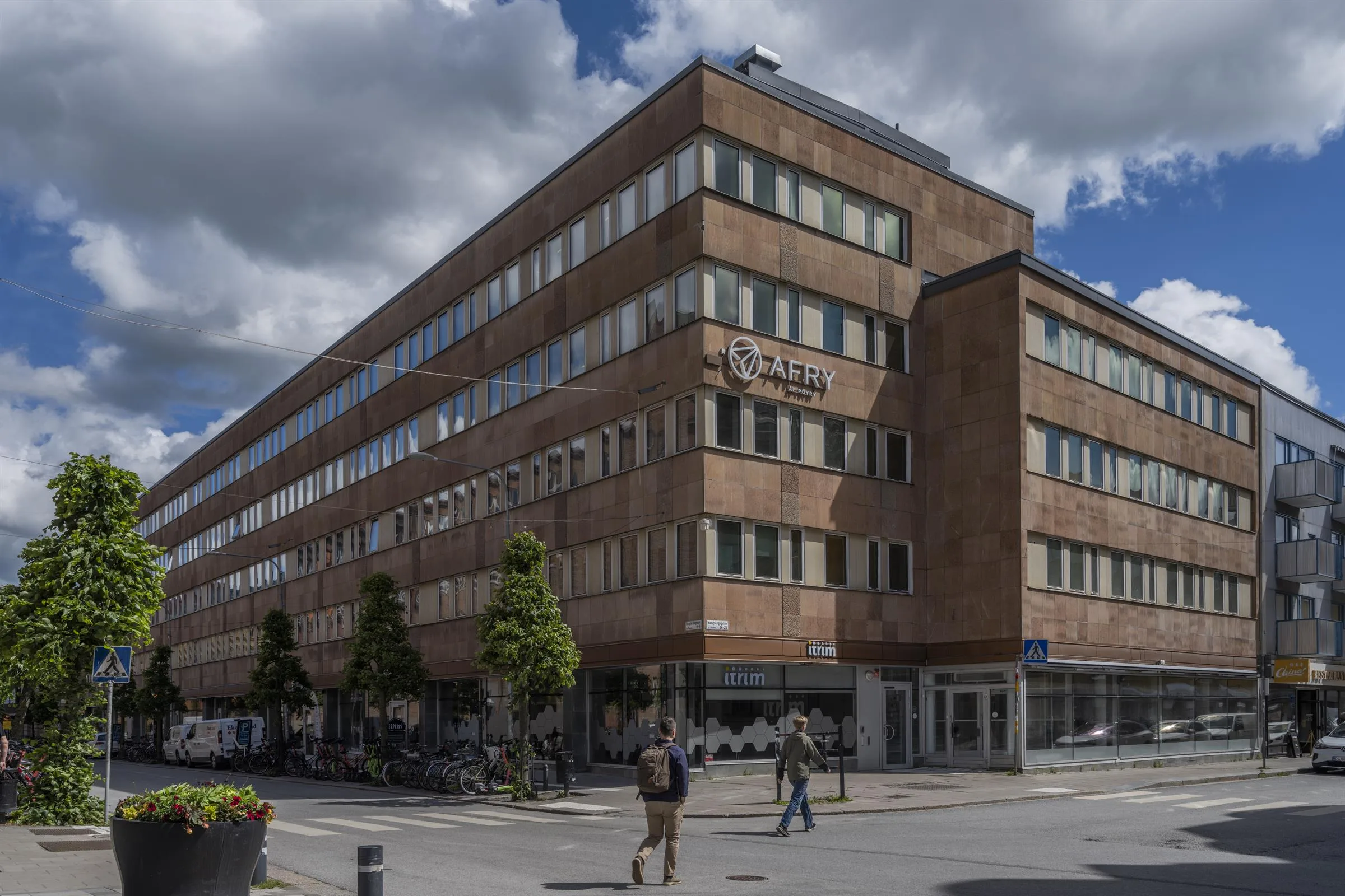 2. En byggnad i tegel med flera våningar, människor går förbi på trottoaren under molnig himmel.