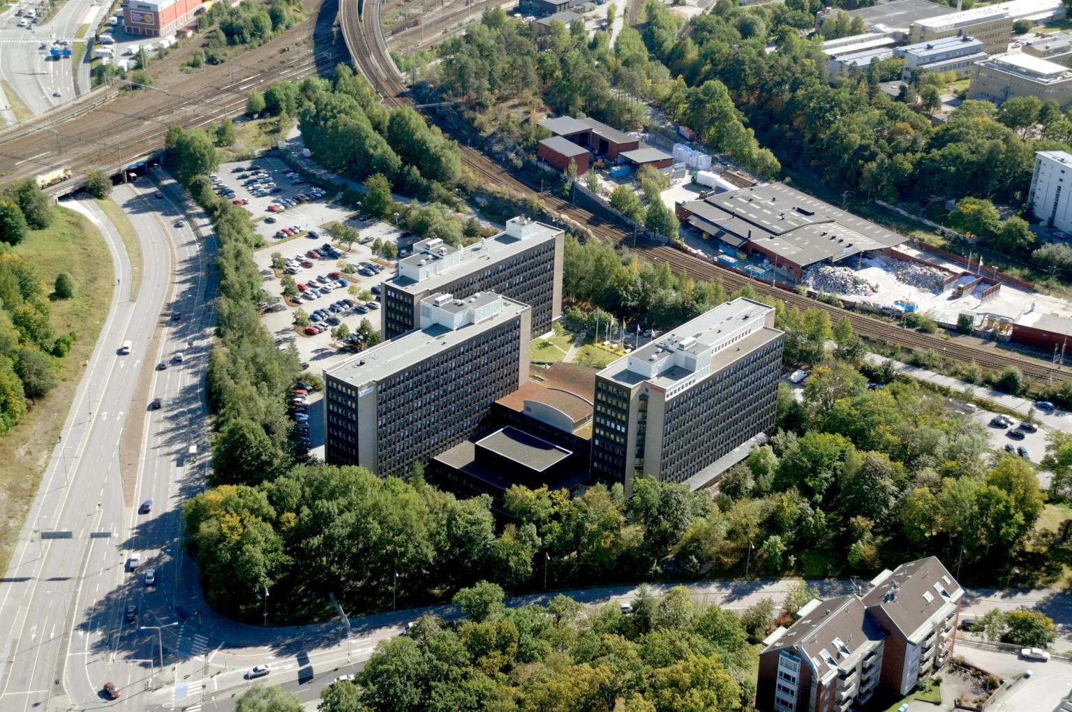 5. Flygfoto över ett område med tre större byggnader omgivna av grönska och parkeringsplatser, med järnväg i bakgrunden.