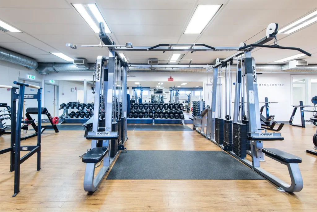 4. Gym med träningsmaskiner, vikter och speglar, samt en ljus interiör med trägolv.