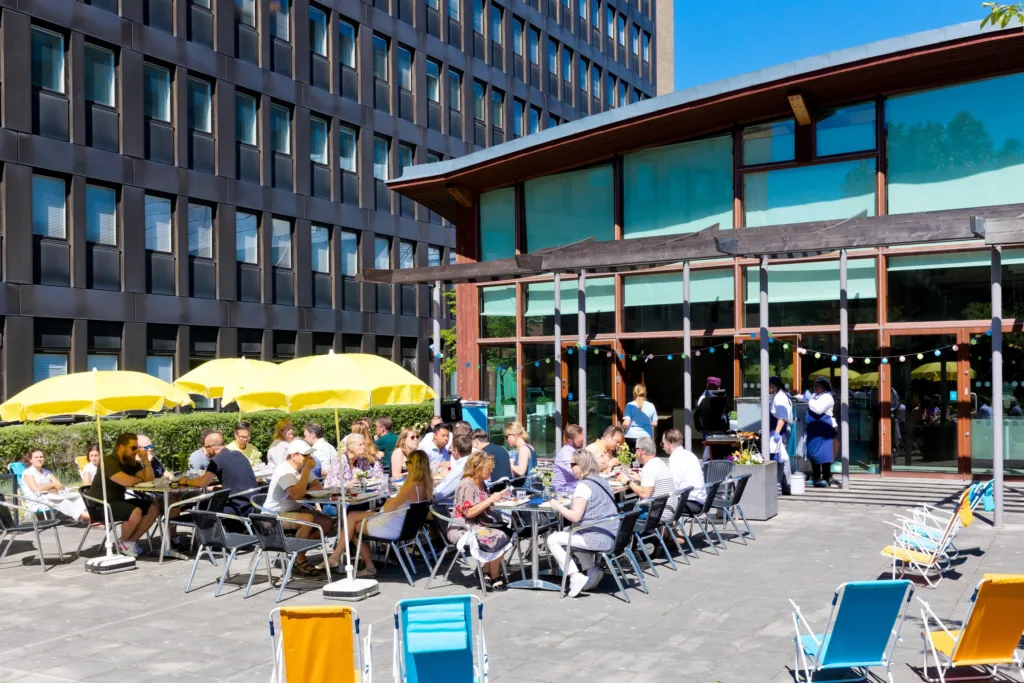 En uteservering med människor som sitter vid bord under gula parasoller, med en modern byggnad och blå himmel i bakgrunden.