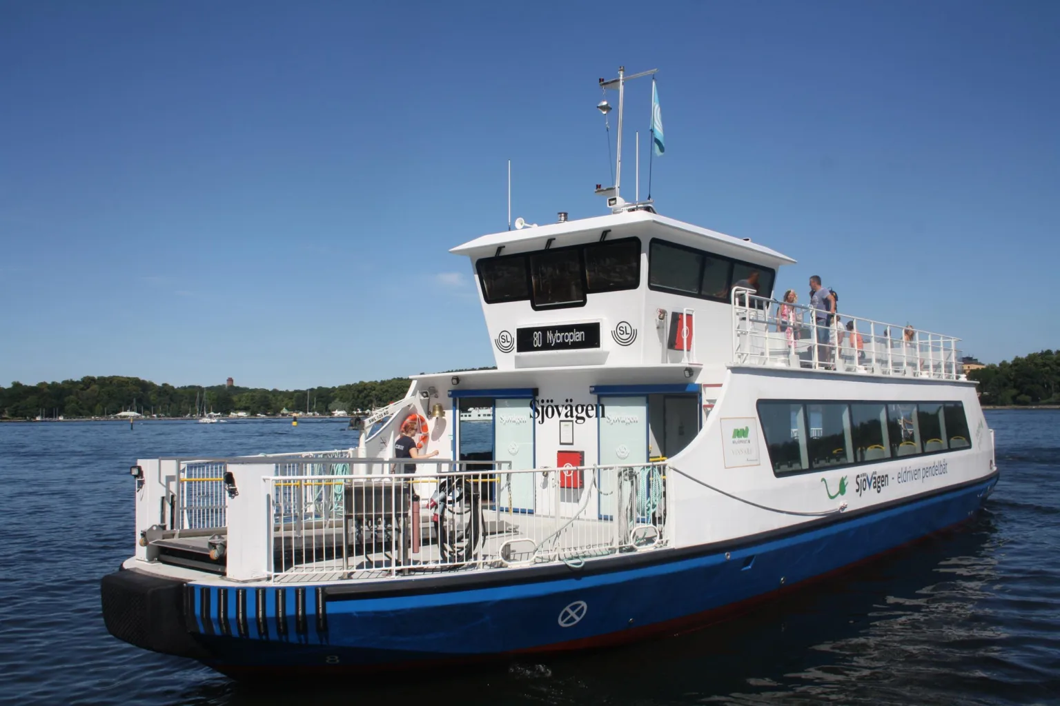 En vit passagerarfärja med blå detaljer ligger vid en kaj, människor står på däck.