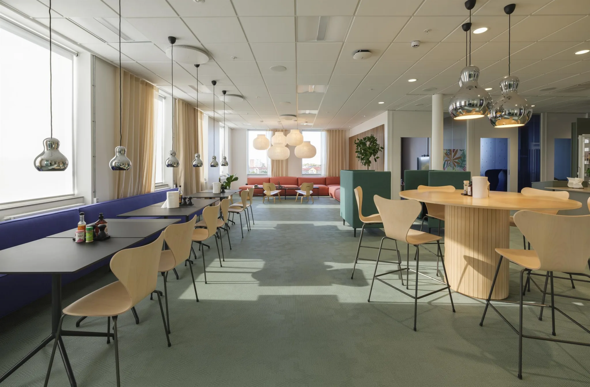 Ett modernt kontorsutrymme med långa bord, stolar och hängande lampor, belyst av naturligt ljus från stora fönster.