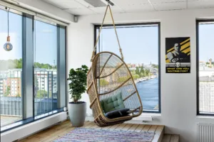 En hängande rottingstol med en grön kudde placerad vid ett stort fönster som vetter mot en stadsmiljö och en flod.