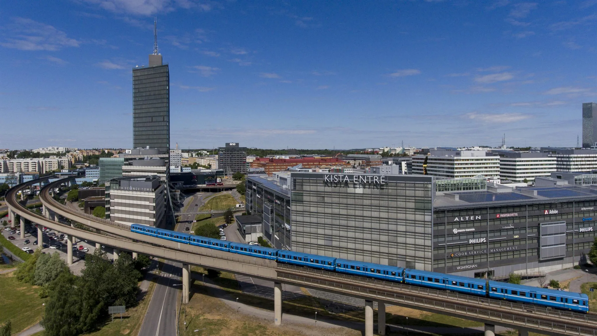 En blå tunnelbanevagn kör på en upphöjd järnväg, omgiven av moderna byggnader under klar himmel.
