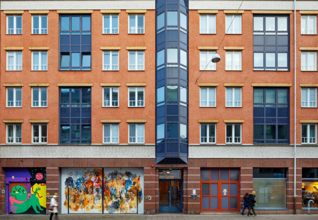 Orangefärgad byggnad med glasdetaljer i mitten, målningar på bottenvåningen och människor som går förbi.