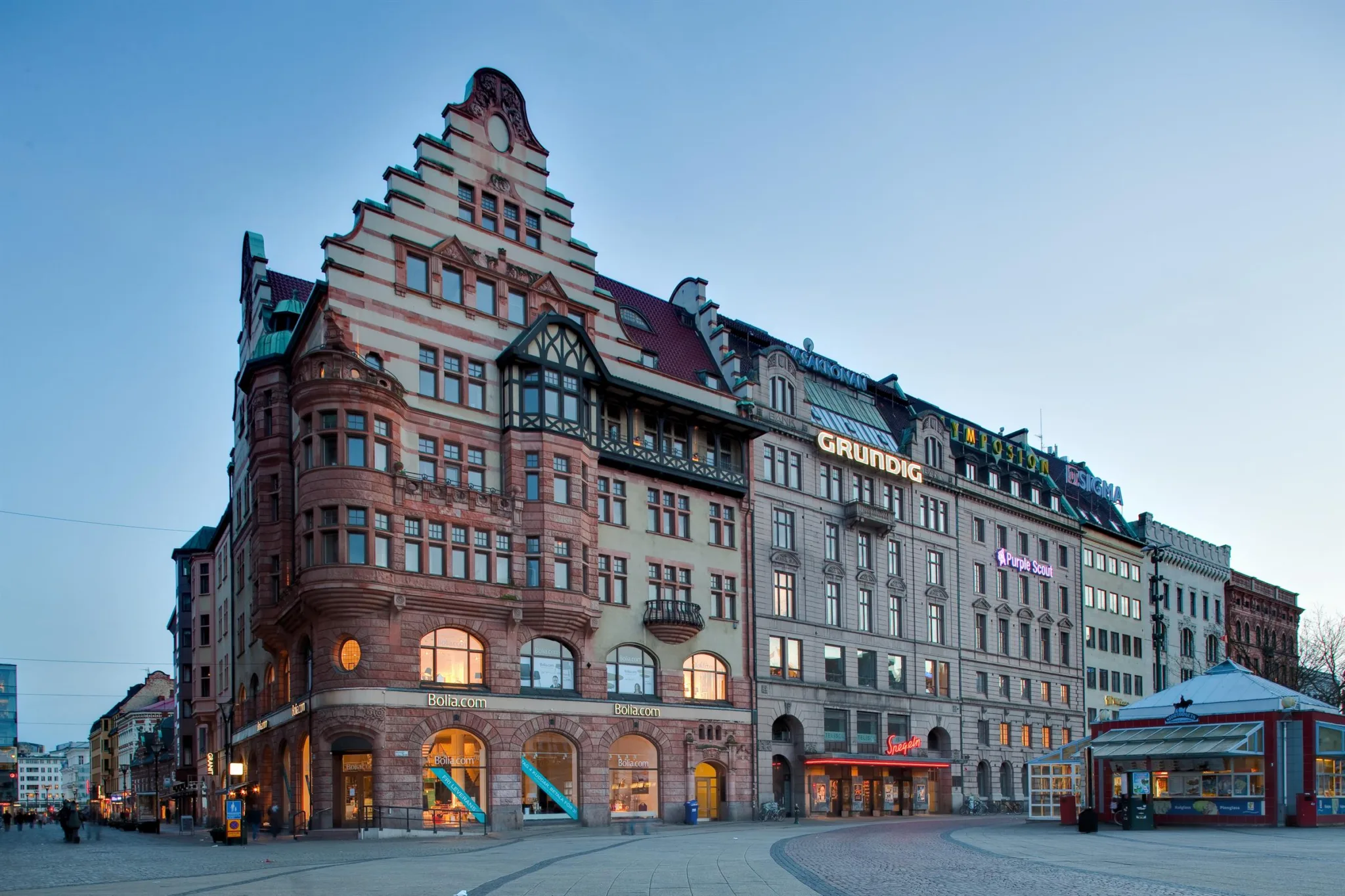 En historisk byggnad med dekorativ fasad och trapptak, omgiven av andra byggnader i stadsmiljö.
