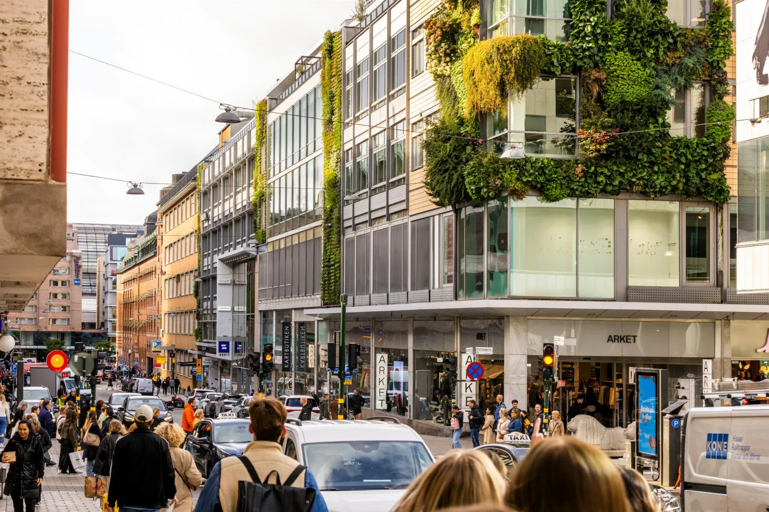 Gatuliv med människor och bilar framför en rad moderna byggnader, varav en har en grön fasad täckt av växter.
