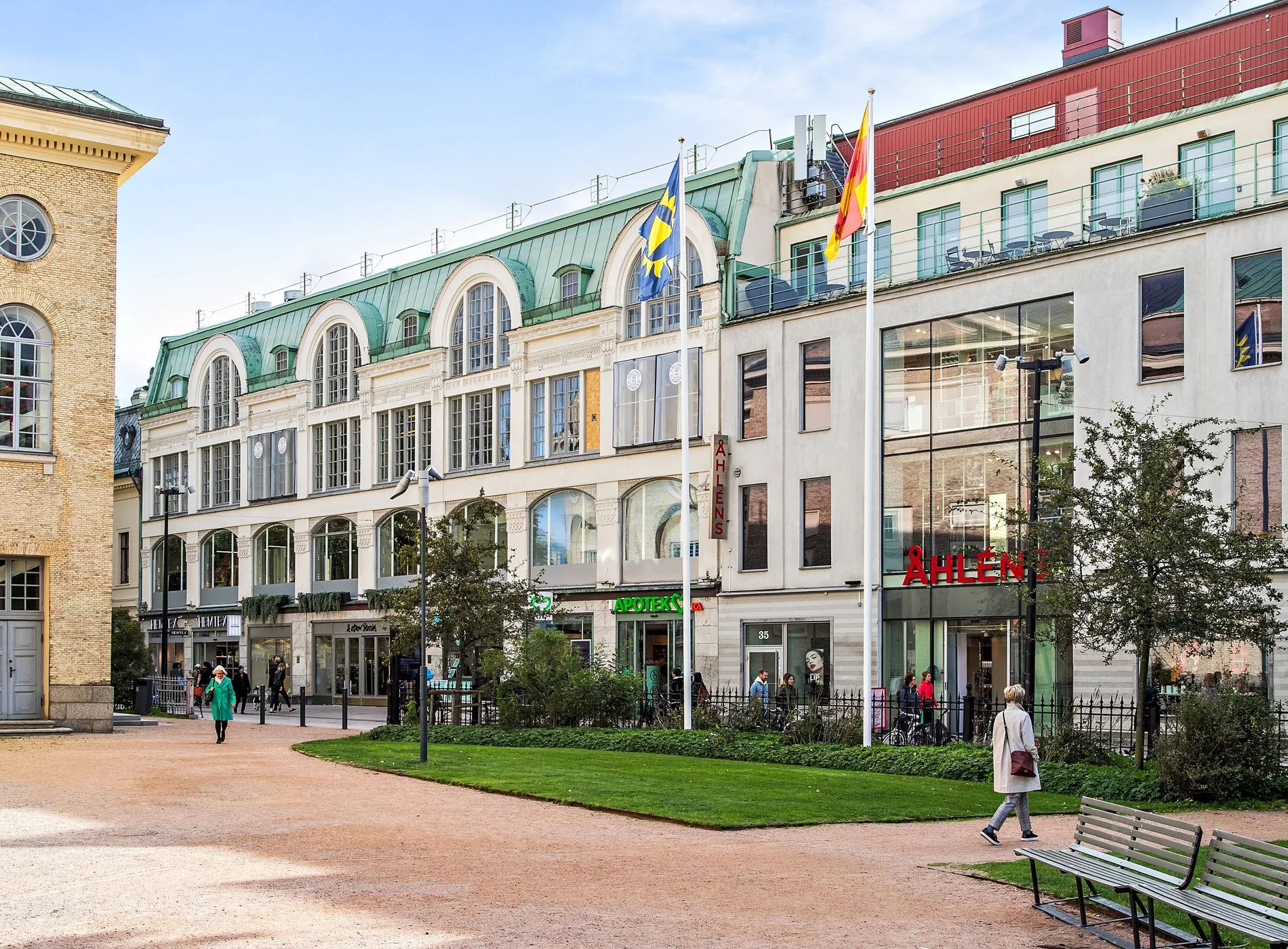En byggnad med stora glasfönster, flaggor och grönska framför, med gångvägar och människor i rörelse.