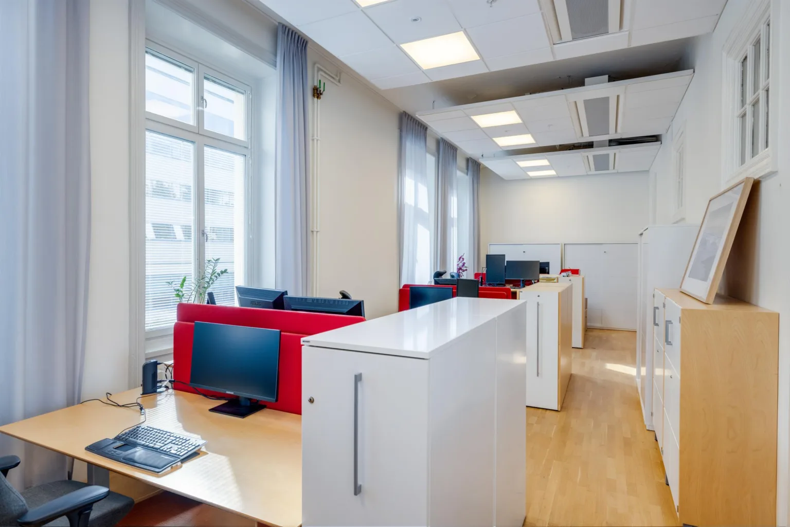 Ett kontorsutrymme med skrivbord, röd inredning, datorer och stora fönster.